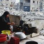 image for A woman cooking food over a small open flame in a heavily damaged urban area in Gaza.